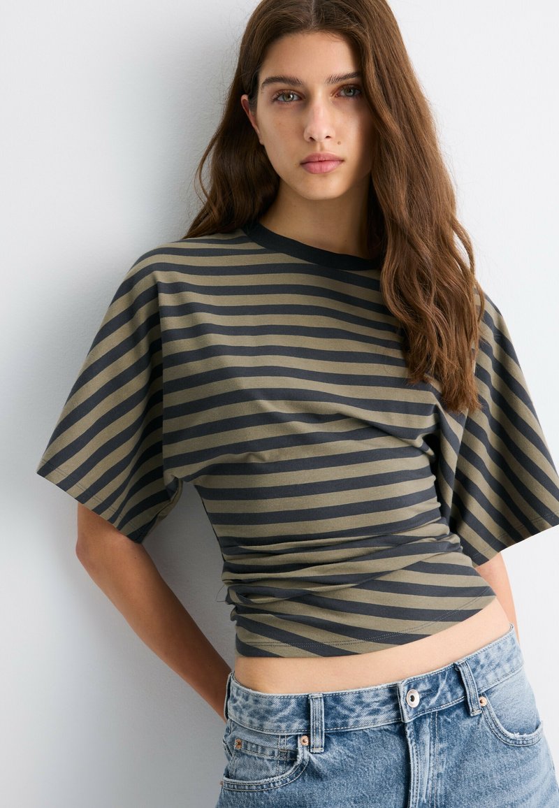 Jeune femme aux longs cheveux bruns portant un haut ample à manches courtes rayé et un jean en denim bleu sur un fond clair.