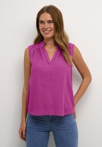 Haut sans manches fuchsia avec une texture côtelée, décolleté en V et détail de volants doux au col, associé à un jean bleu ajusté.