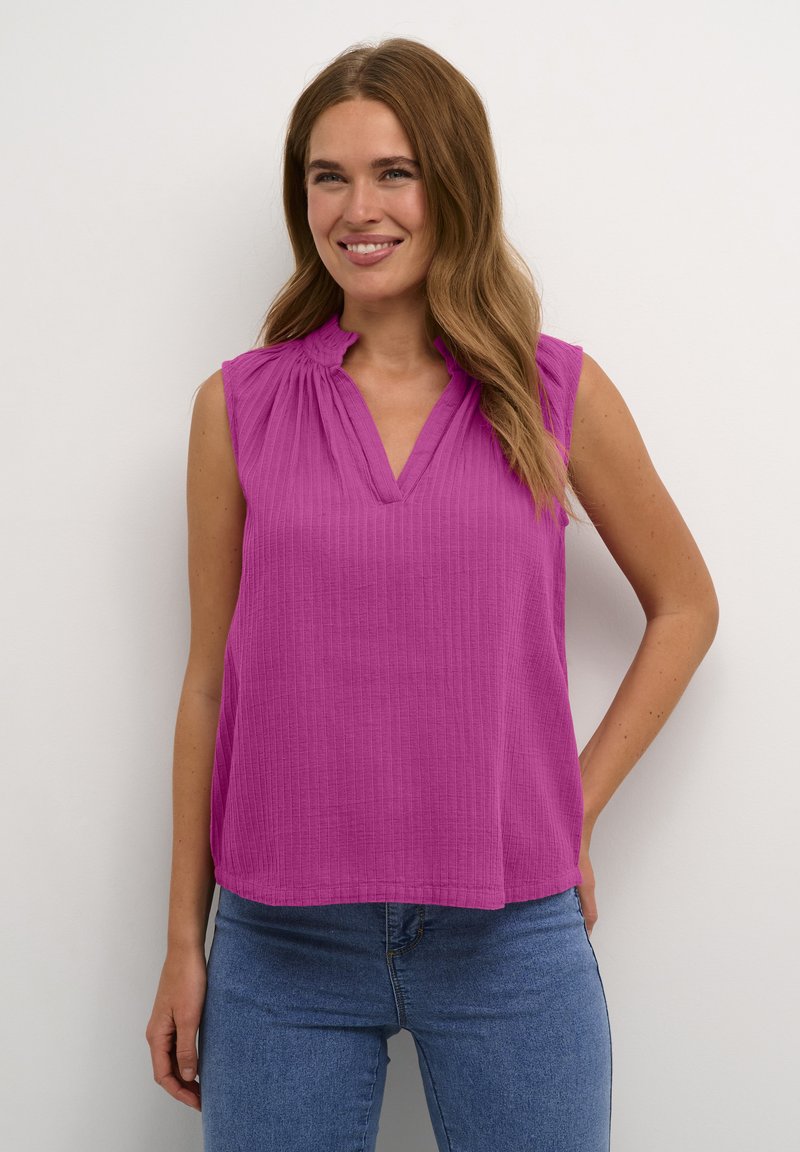 Haut sans manches fuchsia avec une texture côtelée, décolleté en V et détail de volants doux au col, associé à un jean bleu ajusté.