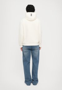 Mann med krøllete hår iført off-white hettegenser og løse blå jeans, stående vendt bort fra kameraet mot en enkel lys bakgrunn.