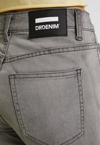 Jeans de mezclilla gris con una textura suave, que presentan una etiqueta de marca negra cosida en la cinturilla y un diseño sencillo de bolsillo trasero.