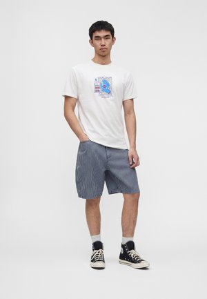 Giovane uomo in piedi con le mani in tasca che indossa una t-shirt bianca con stampa grafica, pantaloncini a righe, calzini bianchi e sneakers nere alte.