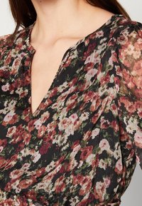 Kukkainen paita, jossa on musta tausta ja punaisia, vaaleanpunaisia sekä vihreitä kukkia, V-pääntie, sekä pehmeä, kevyt kangas, jossa on poimutuksia.