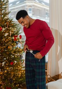 Rood shirt met lange mouwen, gecombineerd met blauwgroene geruite pyjamabroek met verstelbare taillekoord. Kerstboom op de achtergrond.