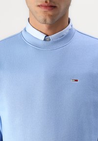 Gaiši zila sporta krekls ar apaļu kakla izgriezumu un ribbētu apkakli, ar mazu izšūtu logotipu ar sarkaniem, baltajiem un ziliem akcentiem.