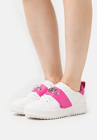Sneakers bianchi con una cinghia rosa, dettagli in argento e una suola testurizzata. Il design include un colletto imbottito e un accent logo sulla cinghia.