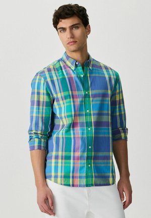 Młody mężczyzna ubrany w koszulę w kratę w kolorach niebieskim, zielonym, żółtym i fioletowym z podwiniętymi rękawami oraz białe spodnie, stoi na jednolitym tle.