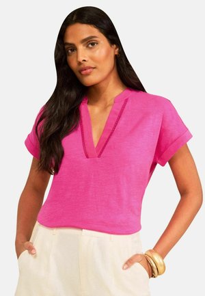 Kvinde med langt mørkt hår iført en lys pink kortærmet top og lyse beige bukser med hænderne i lommerne og gyldne armbånd på håndleddet.