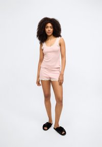 Růžová bavlněná sada pyžama s krajkovými detaily, obsahující tílko bez rukávů a šortky s krajkovým lemováním. Modelka má na nohou černé chlupaté pantofle.