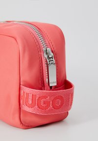 Koralna najlon torbica za kozmetiku s srebrnim zatvaračem i izvezenom trakom "HUGO". Pravokutnog oblika, glatke teksture i minimalističkog dizajna.