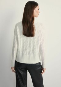 Biały sweter o splocie warkoczowym z ribbowanym wykończeniem u dołu i na mankietach, z teksturowanymi detalami. Noszony na czarnych skórzanych spodniach, podkreślający kontrast całego stroju.