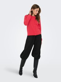 Czerwony kardigan na guziki, zestawiony z czarnymi, luźnymi spodniami i czarnymi botkami na obcasie. Gładka tekstura i swobodny krój.