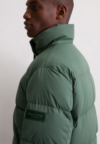 Green puffer jacket with a high collar, quilted design, and a rubber brand patch on the left side. Smooth texture and structured shape.