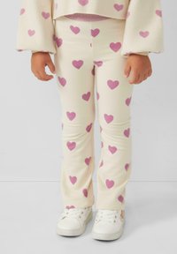 Leggings color crema con una vestibilità aderente, caratterizzati da un motivo di cuori rosa sparsi. Indossati con sneakers bianche per creare contrasto.