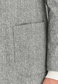 Szaro-skalniasty materiał w wzór jodełki z naszywaną kieszenią. Tekstura wydaje się gładka i miękka, z kontrastowym białym mankietem na krawędzi rękawa.