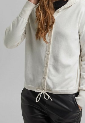 Mujer con sudadera blanca con botones y cordón, y pantalones negros, sujetándose el cabello con una mano cerca de la cabeza.