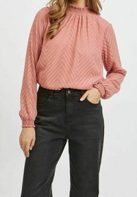 Blusa rosa polveroso con collo alto e arricciato e maniche lunghe, caratterizzata da un motivo a zigzag testurizzato, abbinata a jeans neri a vita alta.