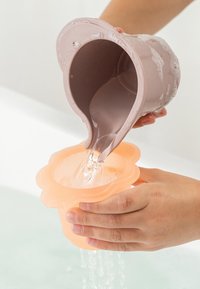 Handen houden een roze, bloemvormige kop vast terwijl er water uit een grotere roze kop in wordt gegoten boven een badkuip.