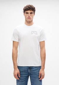 Tricou alb din bumbac cu un mic logo negru imprimat și text în zona stângă a pieptului. Design cu gât rotund și mâneci scurte.