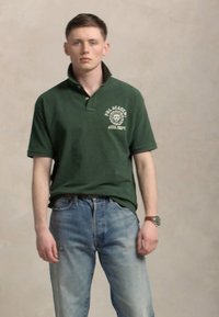 Zelena polo majica s ovratnikom, s logotipom na lijevoj strani prsnog koša, uparen s svijetloplavim trapericama. Model stoji protiv neutralne pozadine.