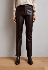 Pantalon en cuir marron avec une coupe slim, doté de poches avant, de passants de ceinture et d'une texture lisse. Porté avec des bottes marron à cheville.