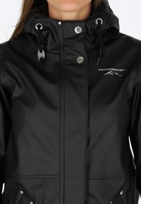 Svart vattentät jacka med hög krage, metalldetaljer, justerbar dragsko och logotyp på vänster bröst. Slät textur och stilren design.