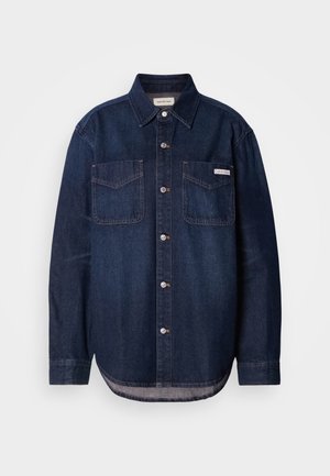 Cămașă din denim bleu închis cu front cu nasturi, două buzunare pe piept și un tiv curbat. Prezintă cusături subtile și etichetă de marcă pe buzunarul din stânga.