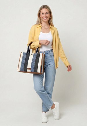 Smiling woman stands wearing yellow shirt, white tank top, light blue jeans, white sneakers, holding large striped tote bag with leather trim.
