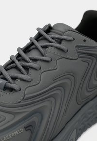 Zapatilla gris con parte superior texturizada con patrones ondulados, cordones planos grises y suela de goma esculpida. Marca sutilmente visible.