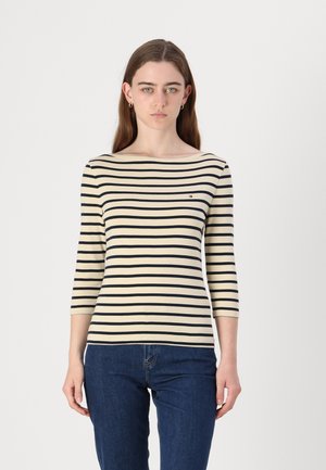 Randig båt-nack tröja i krämfärgad och marinblått tyg, trekvartslånga ärmar, med en liten logotyp på bröstet. Bärs med blå jeans.