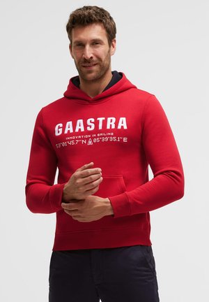 Man wearing red Gaastra hoodie with white text, standing and clasping hands against plain light background.