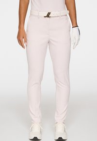 Donna che indossa pantaloni slim-fit rosa chiaro con una cintura abbinata, sneakers bianche e un guanto da golf bianco sulla mano destra.