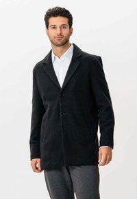 Mand iført sort knapjakke over hvid skjorte og grå bukser, stående foran en helt hvid baggrund.