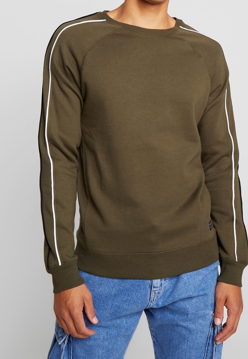 Homme portant un sweat-shirt vert olive avec des rayures blanches sur les manches et un jean cargo bleu, debout devant un fond gris.