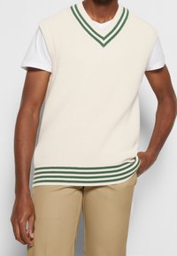 Cremefarbene, gestrickte V-Ausschnitt-Weste mit grünen und weißen Streifen am Kragen und Saum, getragen über einem weißen T-Shirt, kombiniert mit beigen Hosen.