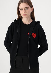 Čierna mikina na zip vyrobená z mäkkého materiálu, so ribovaným spodným lemom a manžetami. Výrazná červená srdcová grafika na ľavej strane hrude.