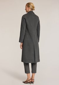 Lungo cappotto grigio in lana con un design strutturato, tasche laterali e una spacco centrale sul retro, indossato sopra pantaloni grigi su misura e scarpe a punta.