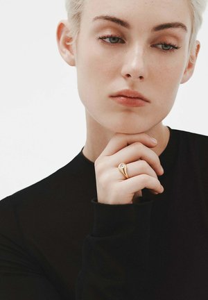 Anillo de oro con una piedra preciosa clara, llevado en un dedo. El modelo tiene el cabello corto y claro y está vestido con una camiseta negra de manga larga ajustada.