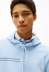 Felpa azzurra chiara con cappuccio regolabile, caratterizzata da un design del logo in bianco e nero sul petto, indossata sopra una camicia a quadri con colletto.