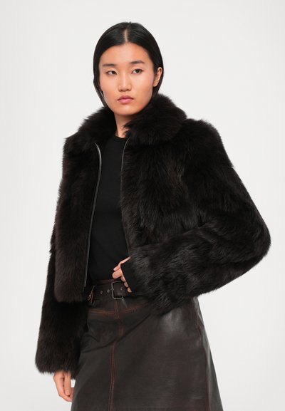 Woman wearing a black fur jacket over a black top and dark brown leather skirt with a belt, standing against a plain white background.
