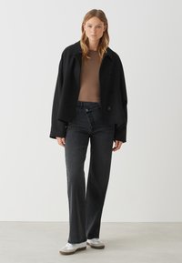 Zwarte, oversized jas met grote revers en knopen, bruine top, high-waisted donkere jeans en witte sneakers met grijze accenten.