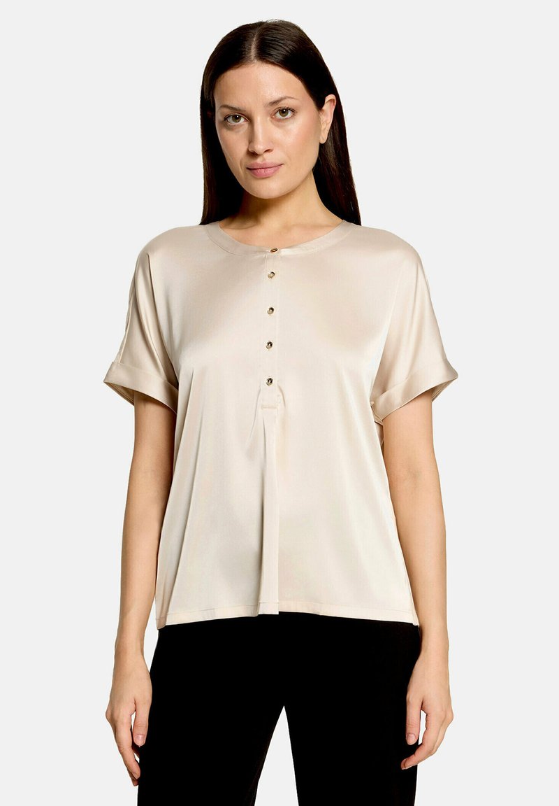 Femme aux longs cheveux foncés porte un chemisier beige à manches courtes boutonné et un pantalon noir, debout devant un fond clair uni.