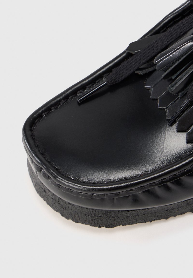 Black leather shoe with a smooth, shiny surface. Features decorative fringe and black laces. Rubber sole with a textured edge.