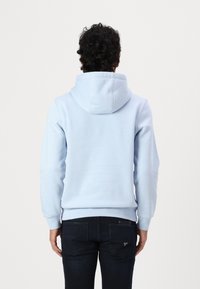Felpa azzurra con un grande cappuccio, polsini e orlo a coste. Il tessuto appare morbido con una superficie liscia. Indossata con jeans scuri.