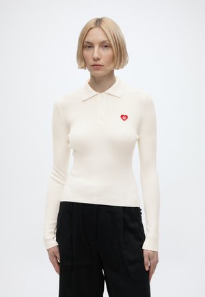 Lyshåret kvinde iført en tætsiddende, langærmet hvid poloshirt med et lille rødt hjerteemblem og løse sorte bukser står foran en hvid baggrund.