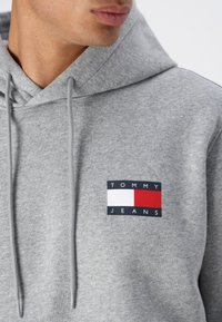 Grå bomulds-hættetrøje med et logo-patch på forsiden, der har sorte, hvide og røde sektioner, samt justerbare snore til hætten.