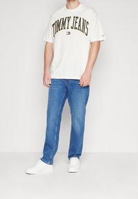 Vit kortärmad t-shirt med "TOMMY JEANS" tryckt i svart, matchad med blå denimjeans och vita sneakers. En enkel design, avslappnad passform.