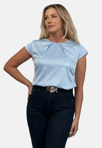 Jasnoniebieska satynowa bluzka z krótkimi rękawami i plisowanym dekoltem, w zestawie z ciemnoniebieskimi dżinsami i czarnym paskiem z metalową klamrą.