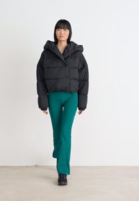 Chaqueta acolchada negra corta con capucha, con una textura acolchada. Combinada con pantalones acampanados verdes y zapatos negros. Fondo blanco liso.