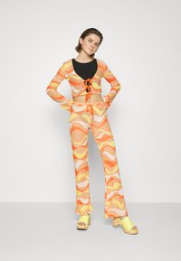 Conjunto de duas peças com uma blusa cropped de manga longa, estampada em laranja, amarelo e branco, combinada com calças largas a condizer. Sabates amarelas.